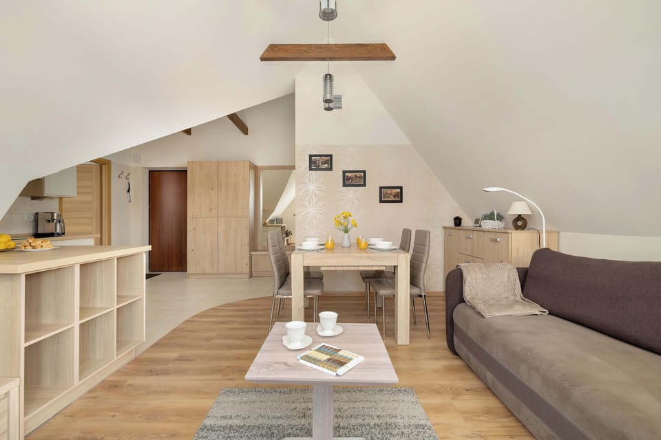 A wide angle showcases the open-plan attic living space, featuring a dining table set for four, a brown sofa, and low shelving units.