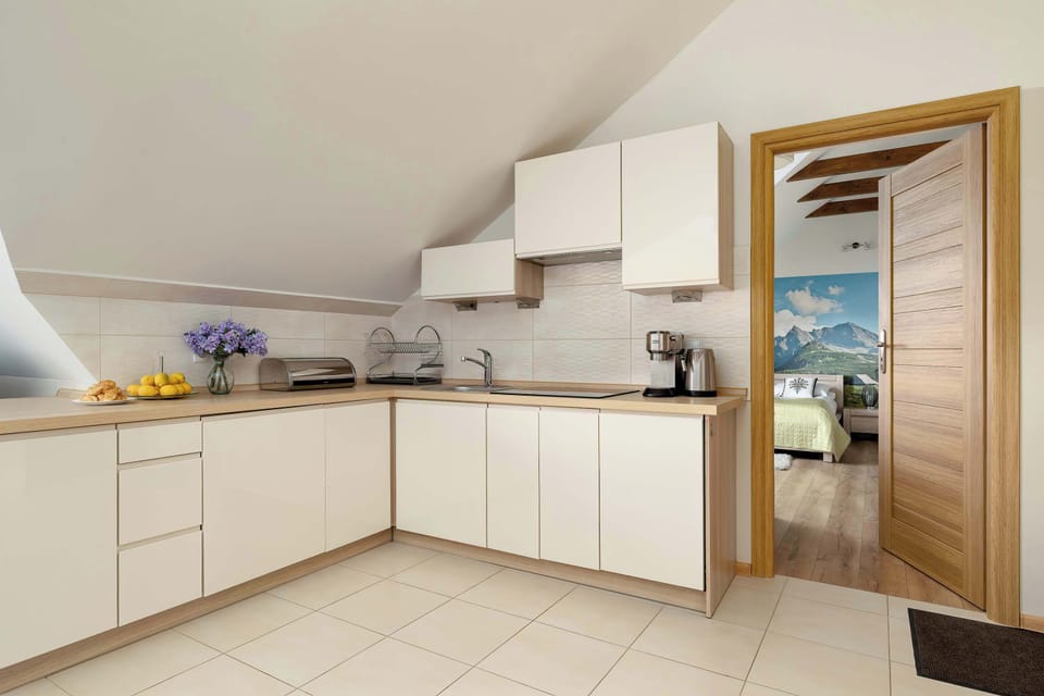 An L-shaped kitchen with white cabinets sits under a sloped attic ceiling. A wooden doorway leads into the adjacent bedroom.