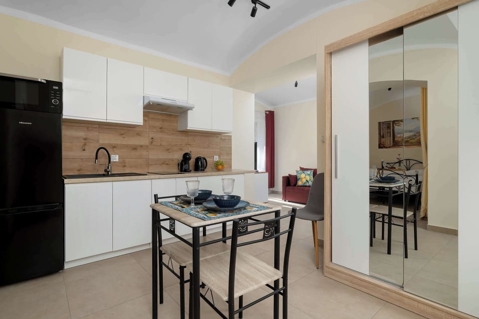 A compact kitchen and dining area with white cabinetry, wooden countertops, and a small table with chairs. The space opens toward the rest of the apartment through a wide archway.