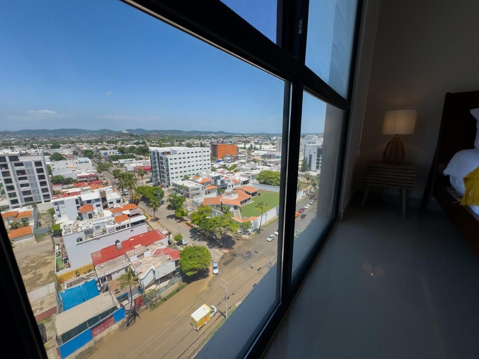 Cityscape view through floor-to-ceiling windows beside a dark wood bed and rattan lamp on tiled floor.