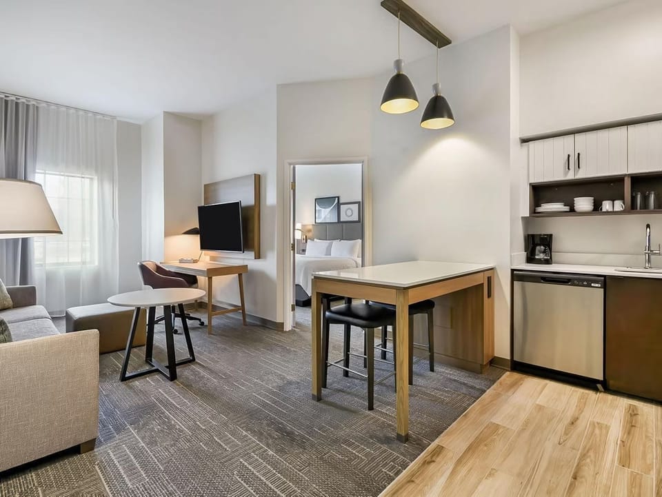 Open-plan living area with a sofa, coffee table, dining table, kitchen, and wall-mounted TV — bright with natural light