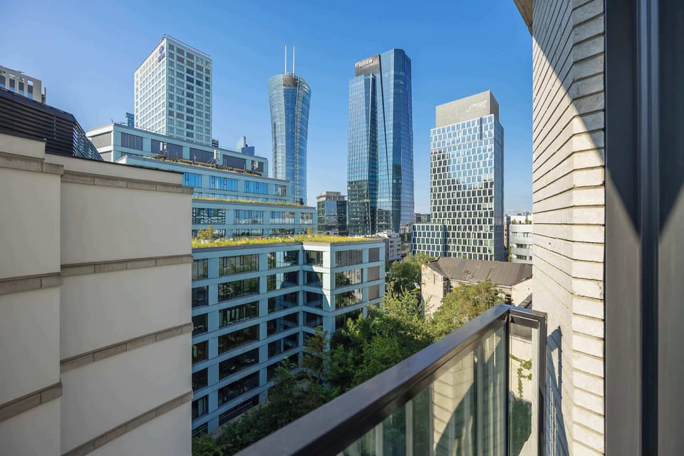 A private balcony offers a direct view of striking city skyscrapers. Metal railings border the space overlooking urban greenery and surrounding architecture. Blue skies brighten the high-rise panorama.