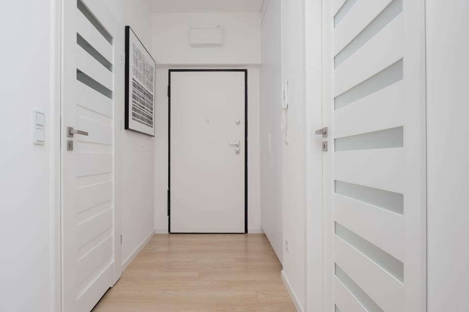 A clean apartment entryway displays light wood flooring and crisp white walls. A sleek front door stands at the end of the hallway between interior doors. Simple geometric wall art adds subtle decoration.