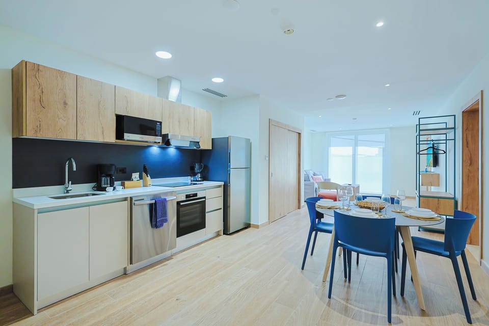 Picture yourself preparing meals in this modern, fully equipped kitchen with sleek countertops, stainless-steel appliances, and ample cabinetry. Enjoy dining at the stylish blue-chaired table before relaxing in the bright, airy living space beyond.