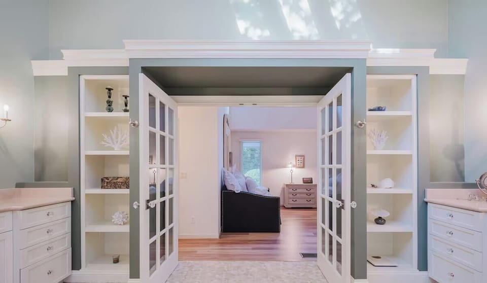 Elegant French doors open to reveal your private bedroom suite, framed by coastal-themed shelving and soft ambient lighting that creates a peaceful, inviting retreat.
