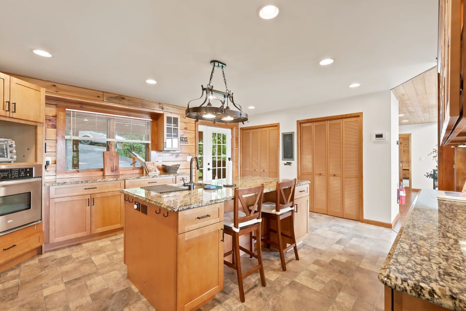 Kitchen with a granite island lined by several wooden bar stools for casual seating, granite countertops, glass-front cabinets, stainless appliances, and a clear sightline through to the great room's wall of windows and layered mountain views beyond.