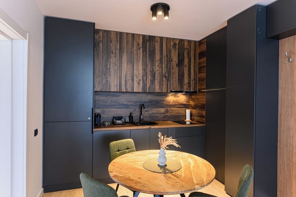 Round solid wood dining table with 3 dark green upholstered chairs, walnut backsplash panelling and anthracite cabinetry. Dining for 3 in the open-plan kitchen of Grand Chalet Suite 1, Poiana Brasov.