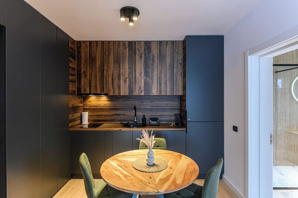 Kitchen and dining area at full view — rustic wood panelling, anthracite cabinetry with under-cabinet lighting, round wood dining table and 3 green chairs on light oak flooring. Grand Chalet Suite 2, Poiana Brasov.