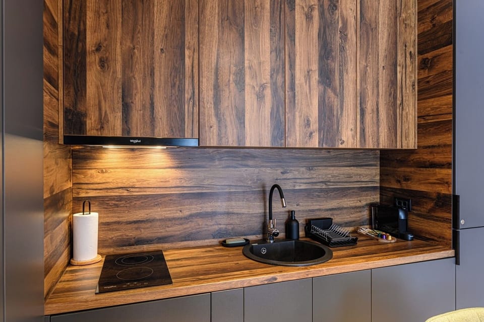 Round black undermount sink with matte black tap set into a solid wood worktop, alongside a 2-ring induction hob. Full-height rustic wood backsplash panelling throughout Grand Chalet Suite 2 kitchen.
