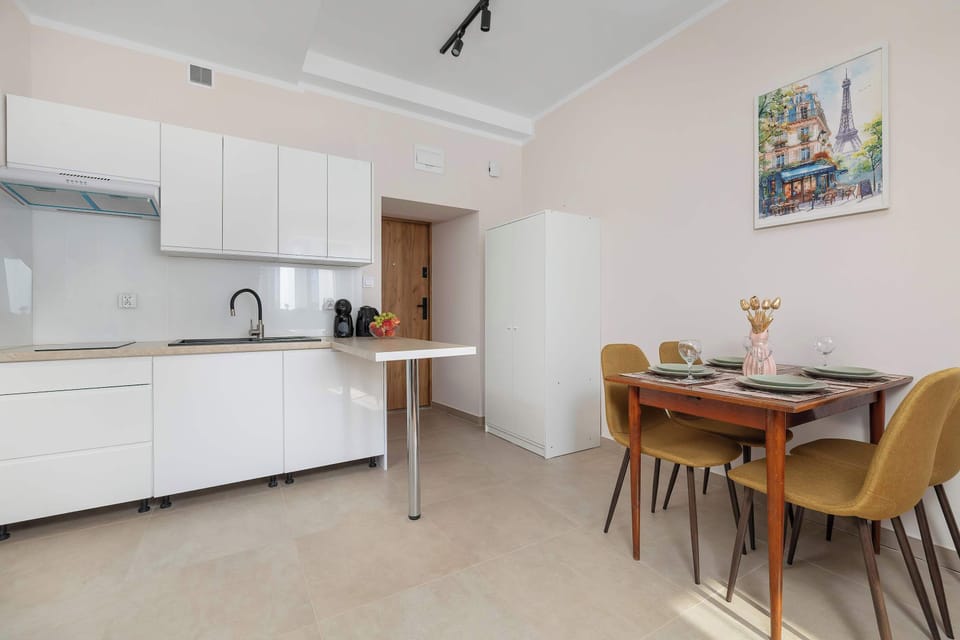 An open kitchenette and dining area features a white kitchenette on one side and a wooden dining table with four upholstered chairs on the other. A colorful painting hangs on the wall above the dining table.