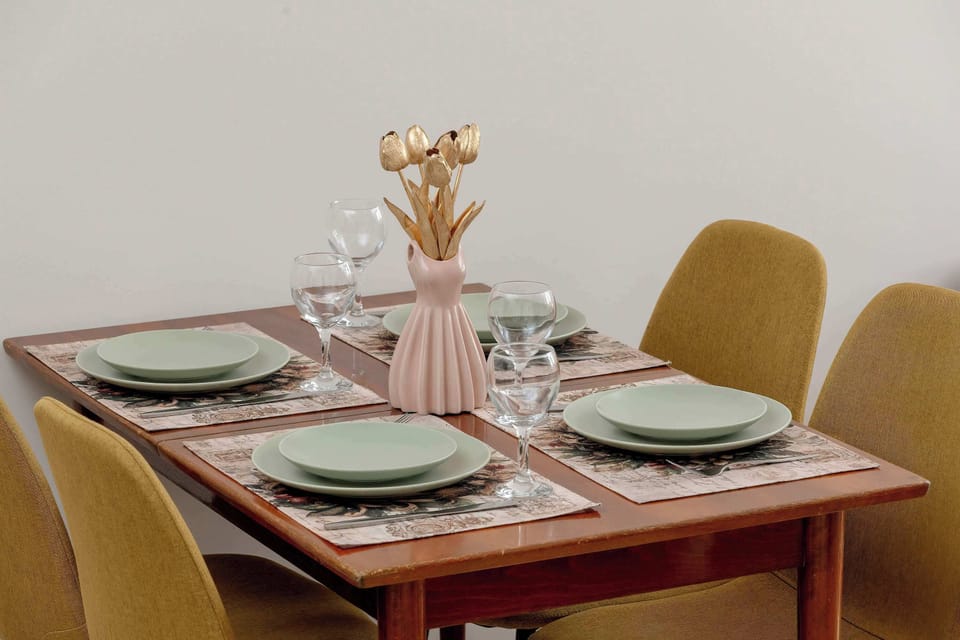 A neatly set dining table is arranged with green plates, cutlery, and glassware, surrounded by comfortable upholstered chairs. A decorative vase with pink flowers serves as a centerpiece.
