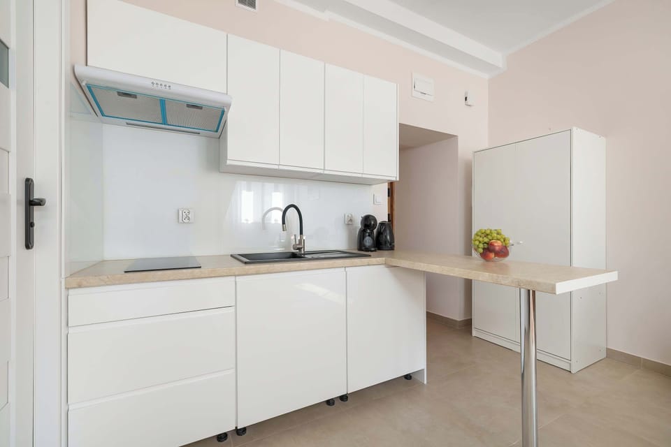 A well-lit kitchennette includes sleek white cabinets, a sink, and a high counter table. A wooden door stands at the end of the short hallway.