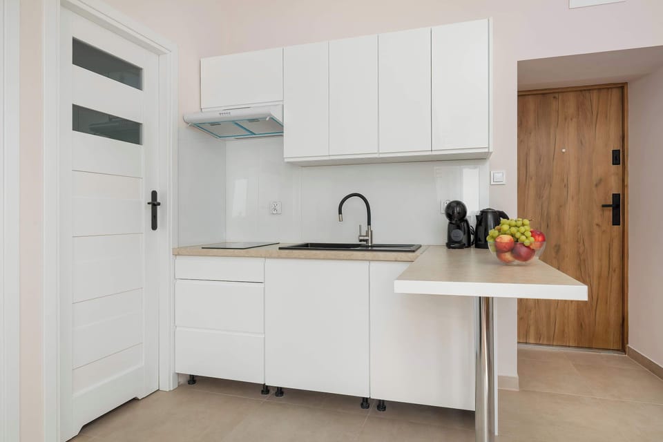 A clean, white kitchenette is visible next to an open doorway, showcasing a minimalist design and a black countertop appliance. The flooring consists of light-colored, glossy tiles.