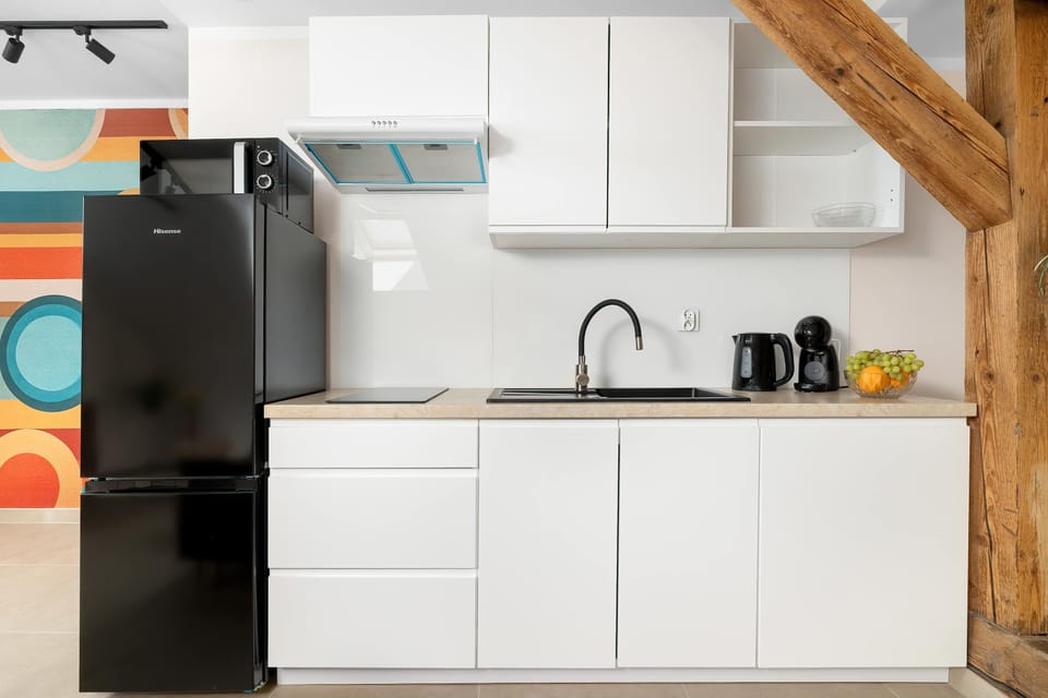 A compact white kitchenette is equipped with a black refrigerator, a sink, and built-in cabinetry. An exposed wooden beam on the right adds a rustic touch to the modern setup.