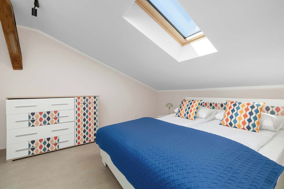 A modern bedroom setup displays a double bed with a blue throw, accompanied by a chest of drawers with a geometric pattern. A skylight sits directly above the bed, offering views of the sky.