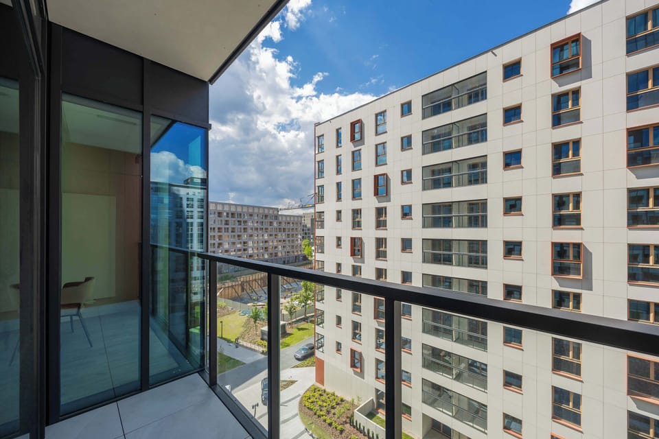 A modern balcony with a glass railing provides a view of adjacent high-rise residential buildings. The clear blue sky and surrounding architecture create a distinct urban feel.