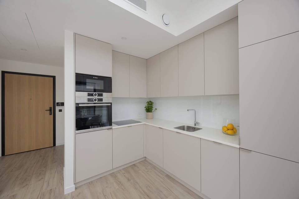 A sleek, modern kitchenette is equipped with minimalist white and light grey cabinets. Built-in appliances and a dark backsplash add a sophisticated contrast to the bright room.