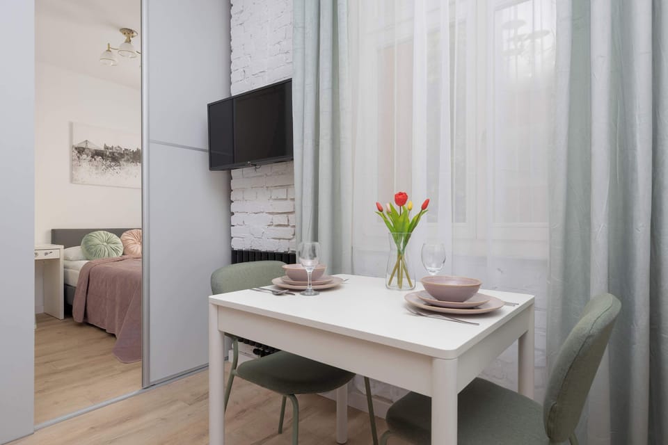 A dining table is placed beside the bed near a large window. A TV is mounted on the wall opposite the bed.