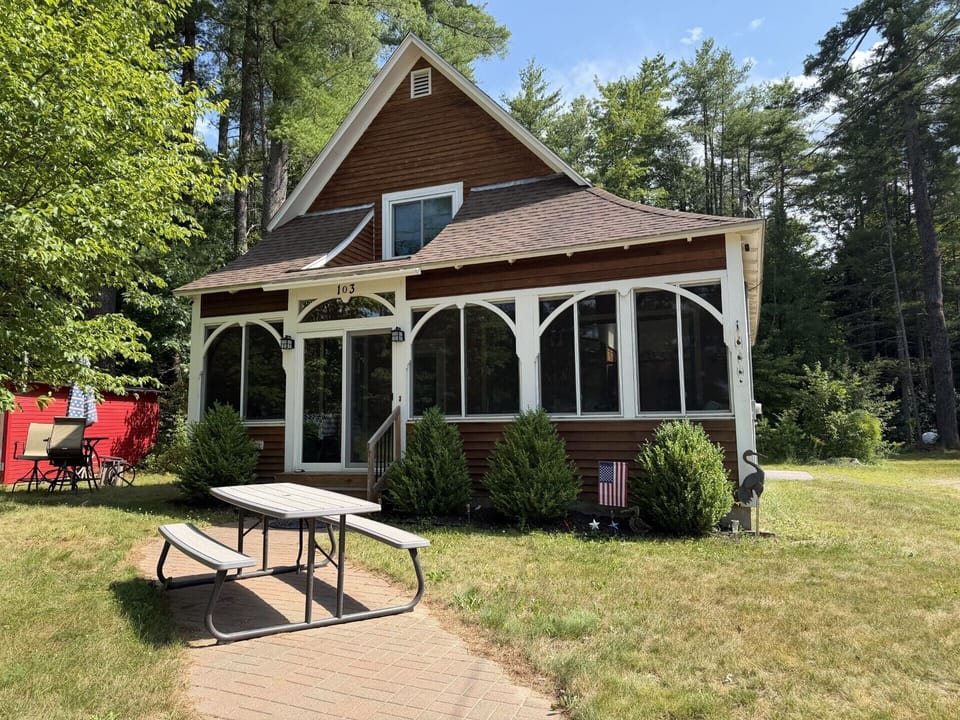 Charming cedar cottage nestled among towering pines, featuring a sunroom with graceful arched windows and a spacious lawn with a picnic table perfect for morning coffee and outdoor dining.