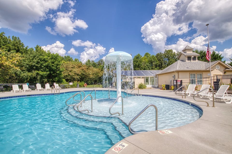 Pool. Clear water. Fountain. Chairs. Green trees. Blue sky. Bushes. Buildings.
