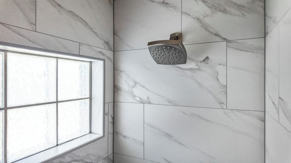 Renovated shower enclosure featuring sleek marble-look tile, a modern showerhead, and natural light from the privacy window.