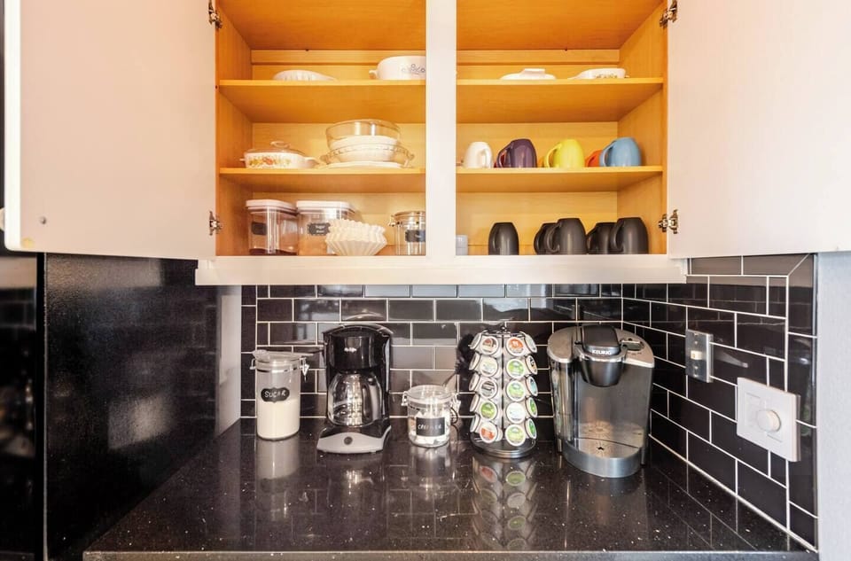 Convenient coffee station with a Keurig, drip coffee maker, and a selection of coffee pods. Cabinets are stocked with mugs, filters, and serving essentials. Black countertops and backsplash provide a sleek, modern look.
