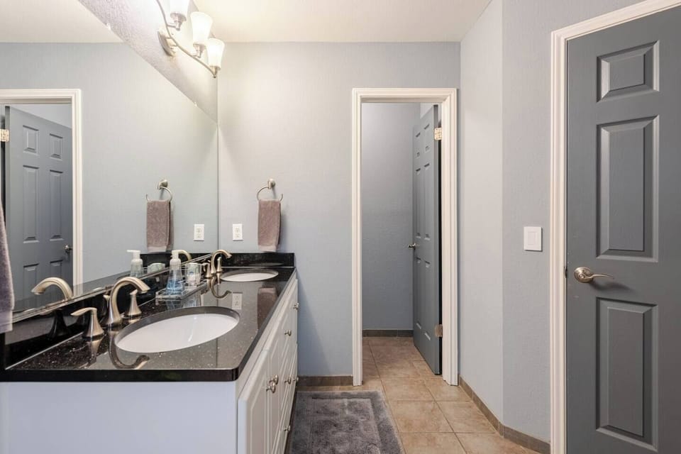 Spacious full bathroom featuring dual sinks with sleek black countertops, ample lighting, and soft gray walls. Includes a private toilet area and a tiled floor for comfort and functionality.