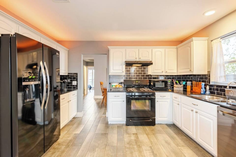Spacious kitchen with black appliances, including a gas stove, microwave, refrigerator, and dishwasher. White cabinets and black tile backsplash add style, while wood-style floors and a sunny window complete the space for cooking and dining.
