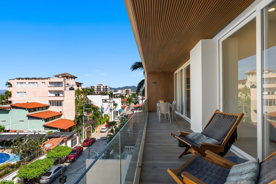Covered private balcony featuring a modern wood-paneled ceiling, glass railings, woven loungers, intimate outdoor dining, and street views via sliding doors.