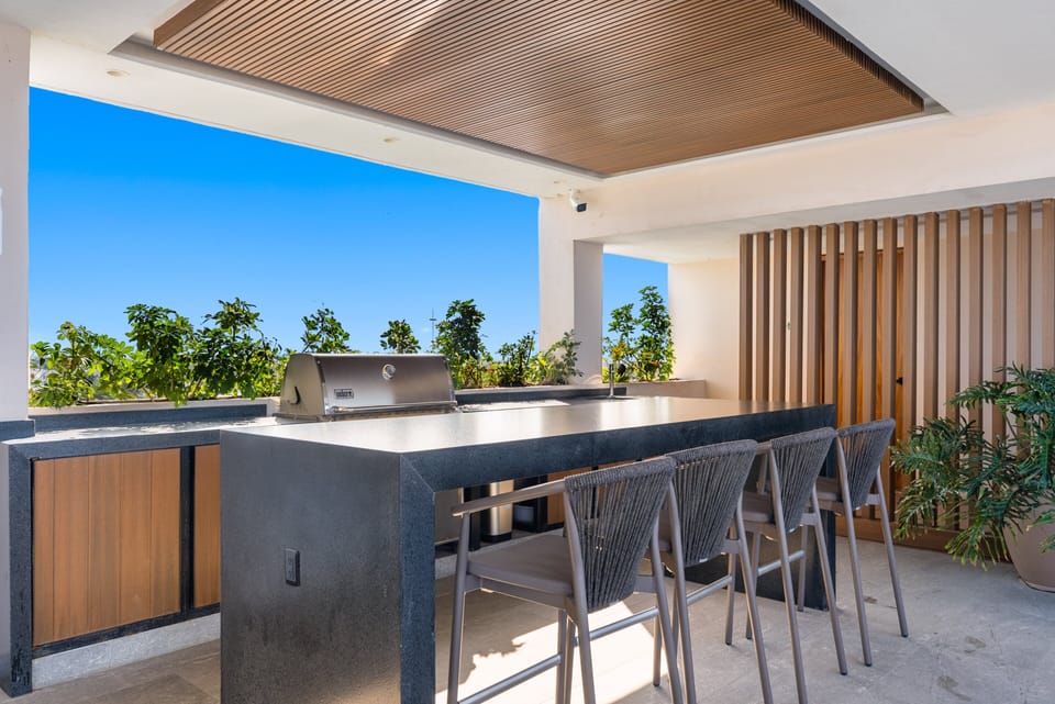 Rooftop outdoor kitchen with a stainless steel grill, sleek black island, woven bar stools, wood-slat ceiling, and coastal greenery under clear skies.