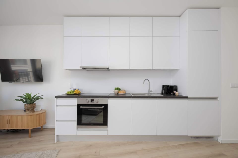 A straight-on view displays the full length of the minimalist white kitchenette. A wooden media console is visible on the far left.
