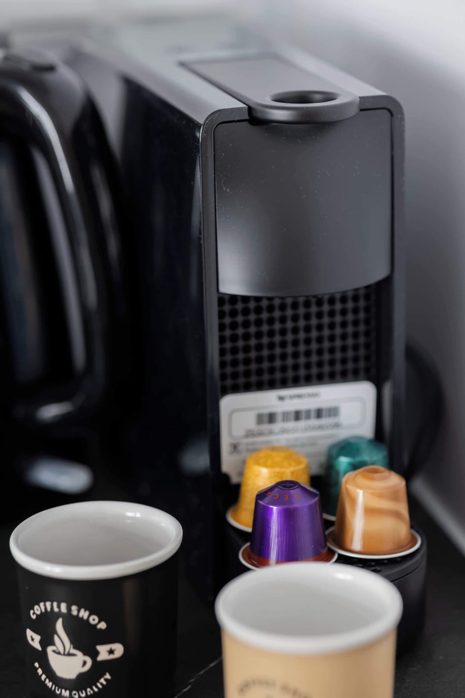A detailed shot focuses on a black capsule coffee machine resting on a counter next to colorful coffee pods and small paper cups.