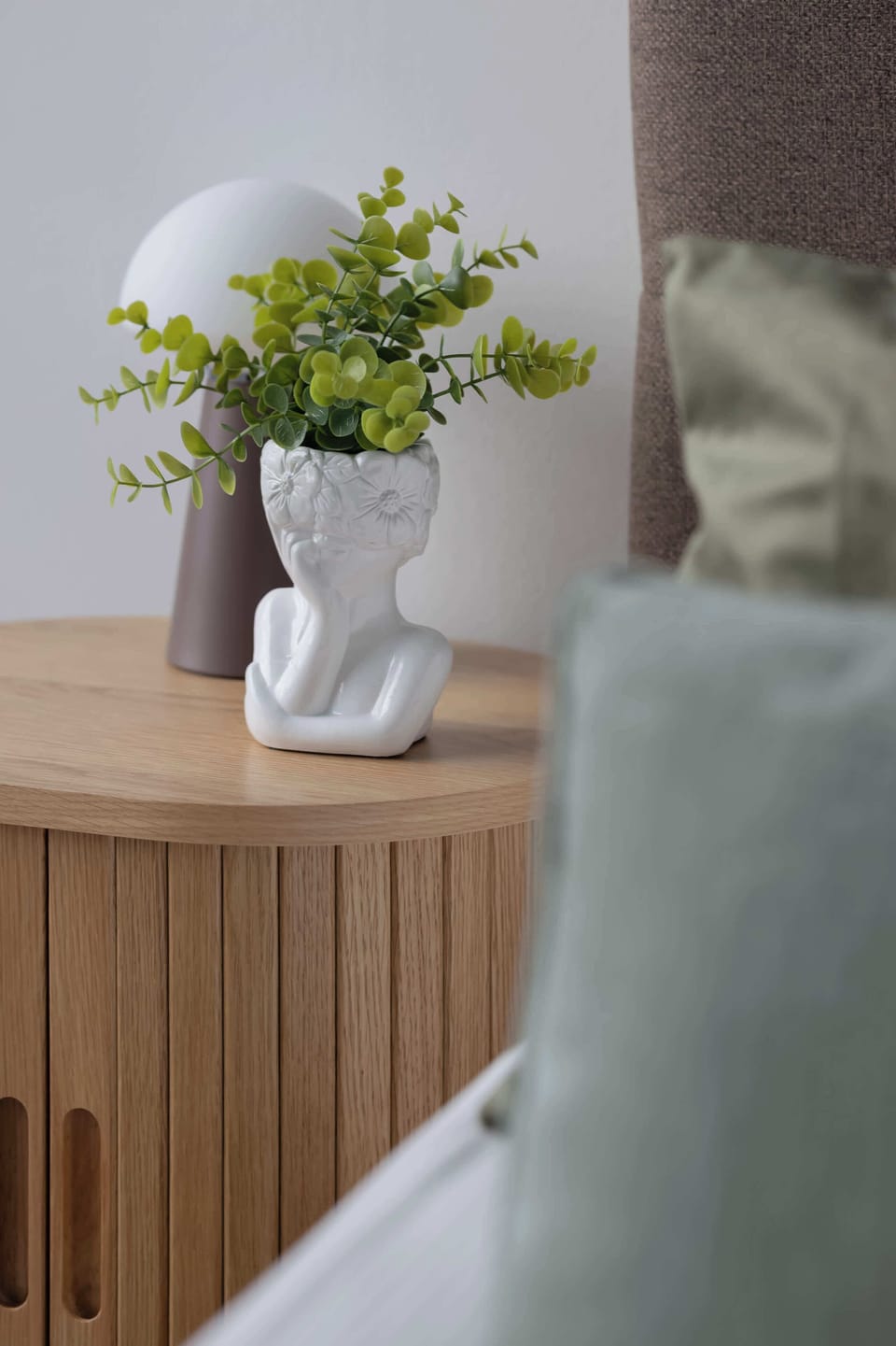 A vertical close-up displays a fluted wooden nightstand holding a small lamp and a unique face-shaped ceramic vase with green foliage.