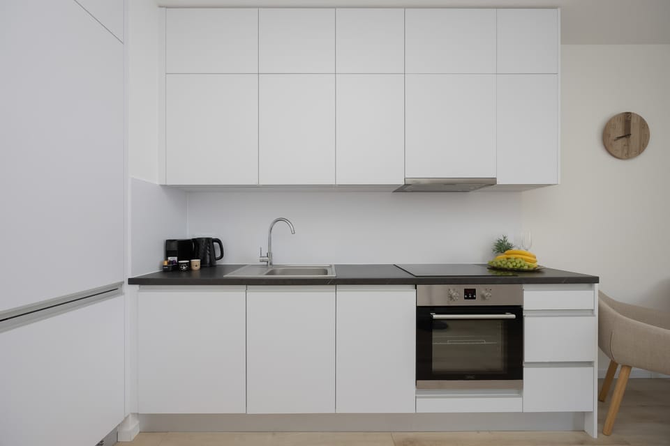 A sleek white linear kitchenette features handleless cabinets, a dark countertop, and a built-in oven. A simple wooden clock decorates the wall.