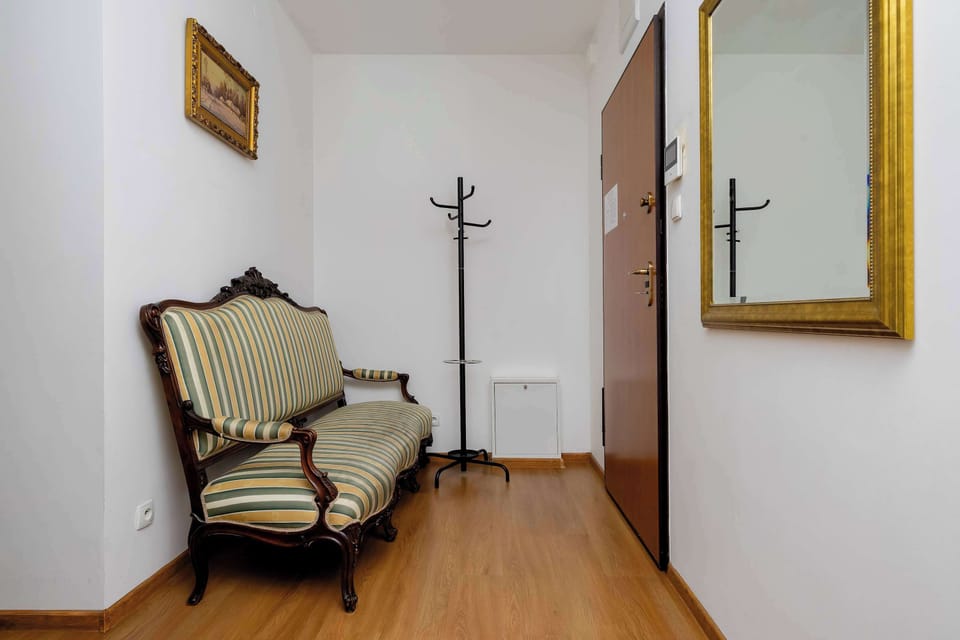 A bright hallway features an elegant, classic bench with patterned upholstery. A tall mirror and a simple standing coat rack stand against the clean white walls.