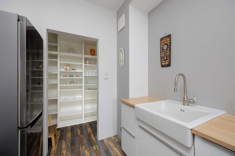 A modern kitchenette space features a white sink set into a wooden countertop. The view extends down a hallway with wooden floors, revealing a large stainless steel refrigerator.