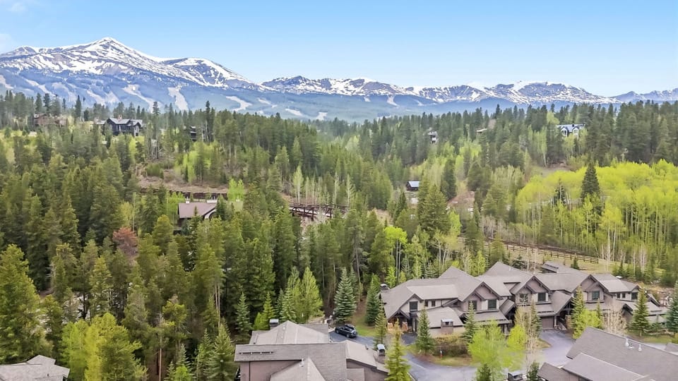 Aerial view of the property nestled among tall evergreens and aspens, with snow-dusted mountain peaks rising majestically in the background. The vacation rentals sit peacefully within a forested mountainside neighborhood.