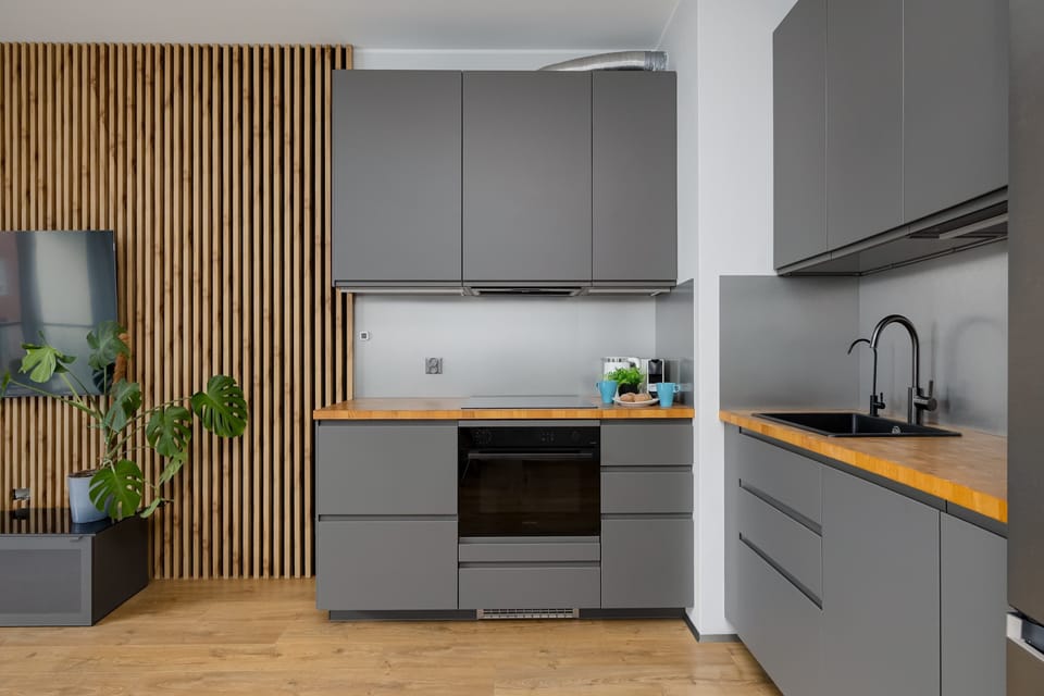 A sleek L-shaped kitchen combines matte grey cabinets with warm wooden countertops. A vertical slatted wooden wall panel sits beside a potted monstera plant.