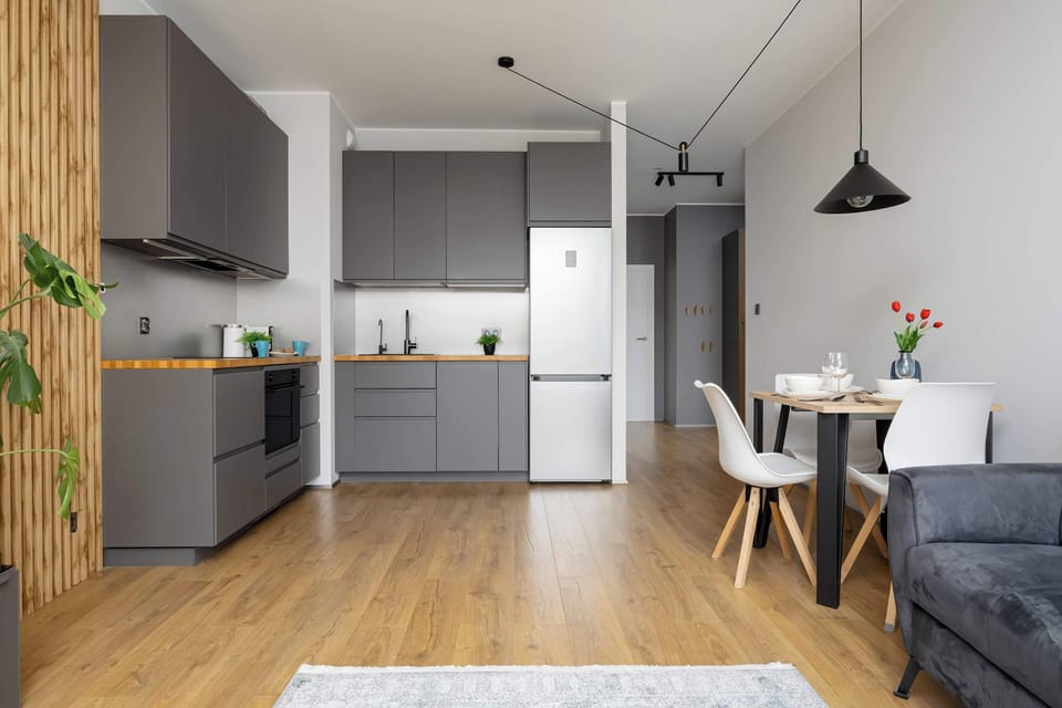 The open layout connects a grey L-shaped kitchen directly to the dining table and entryway. White walls and wooden countertops create a clean contrast.