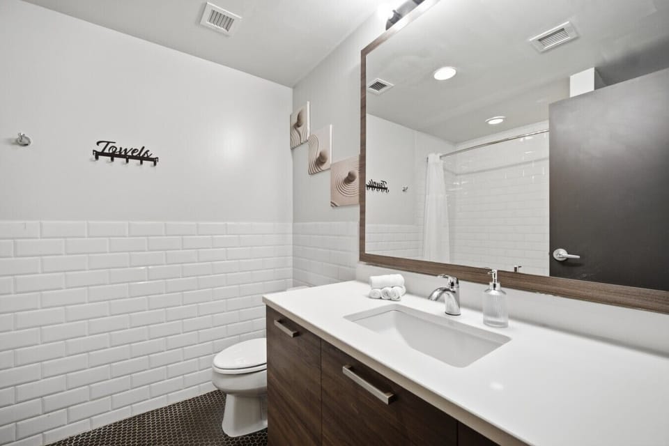 Bright apartment bathroom featuring a vanity with a white countertop and sink, a large mirror, white subway tile walls, a toilet, and a towel hook rack.