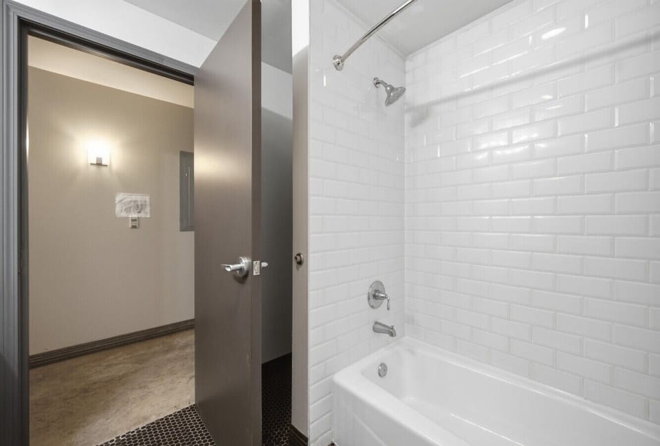 Clear apartment bathroom featuring a white bathtub with tile surround, a shower head, and an open doorway leading to the hallway.
