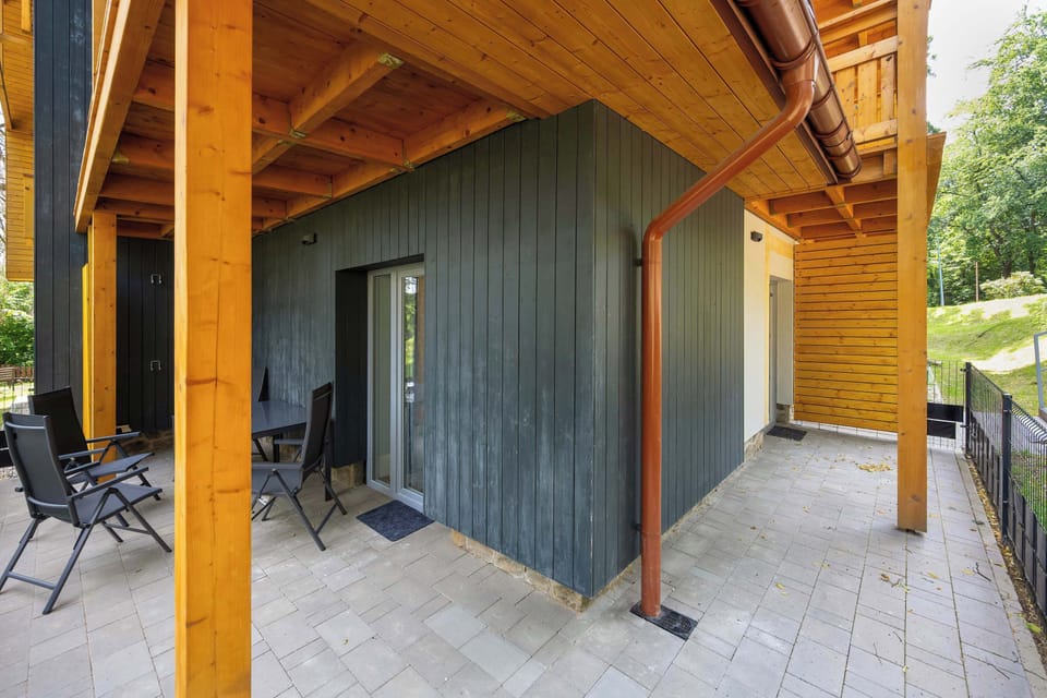 A ground-level terrace features a seating area sheltered by a wooden overhang and a yellow pillar. The outdoor space is beautifully framed by natural scenery and modern architecture.