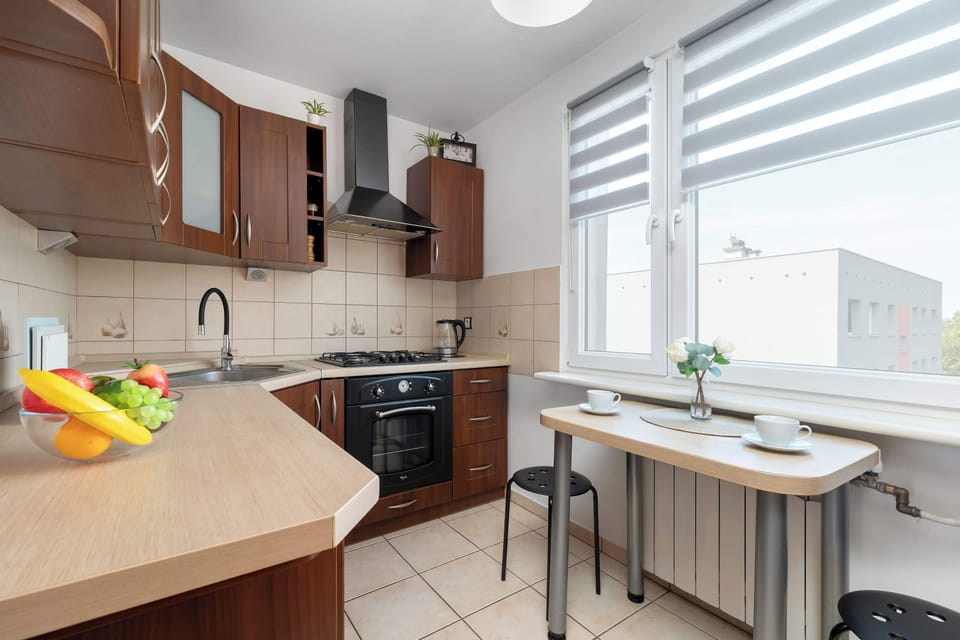 A view of the kitchen corner showing a compact dining table set for two next to a window with roller blinds. Wooden cabinets provide ample storage space along the wall.