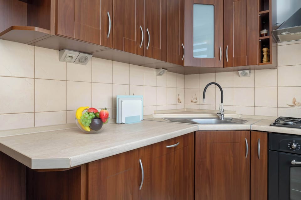 A close-up angle of the L-shaped kitchen featuring wood-finish cabinetry, light stone-effect countertops, and a stainless steel sink. Under-cabinet lighting illuminates the prep counter.