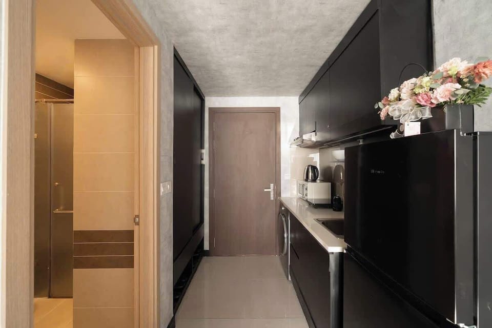 View of the streamlined galley kitchen with sleek black cabinetry and light countertops. This angle shows the apartment door ahead and the bathroom entrance on the left, highlighting the highly efficient layout.