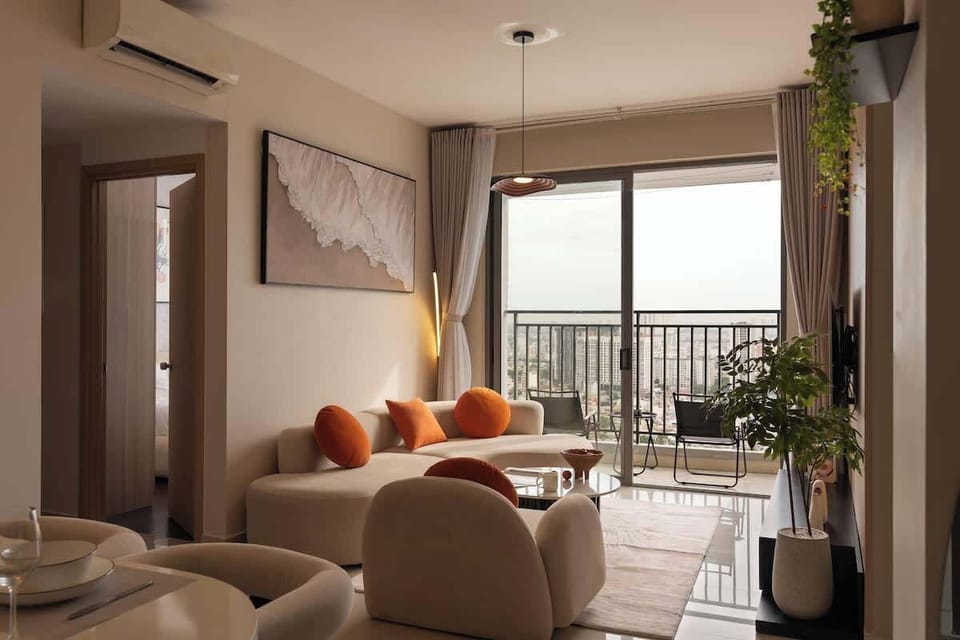 The living room features cozy seating, warm decor, and a smart TV. Floor-to-ceiling glass doors lead to a balcony with stunning river views. Natural light elegant accents, and greenery create a bright and relaxing space for unwinding or entertaining