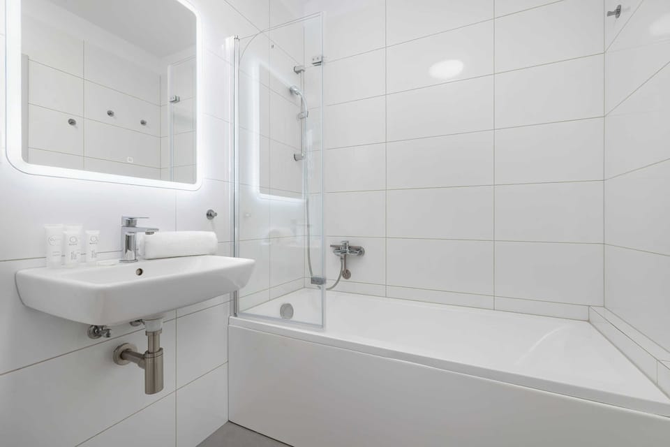 A clean, modern bathroom is equipped with a white bathtub and a clear glass shower screen. An illuminated mirror hangs above the sink, set against bright white tiled walls.