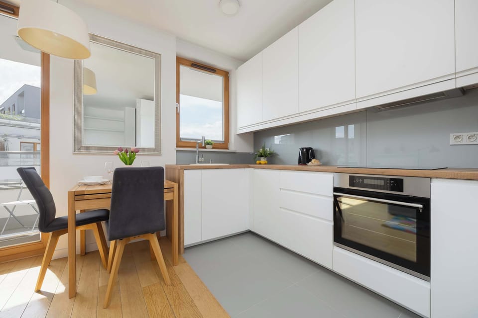 A bright kitchen corner with white cabinetry, wooden flooring, and a small dining table placed beside the window.