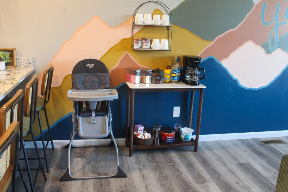 A cheerful coffee and snack station awaits you with a high chair ready for little ones, all set against a vibrant mountain-themed mural.