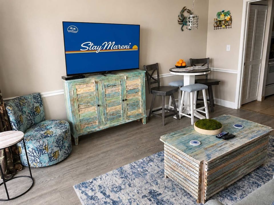 Bright, coastal-inspired living room with a large Smart TV, cozy seating, and a charming dining nook perfect for relaxing after a day at the beach.